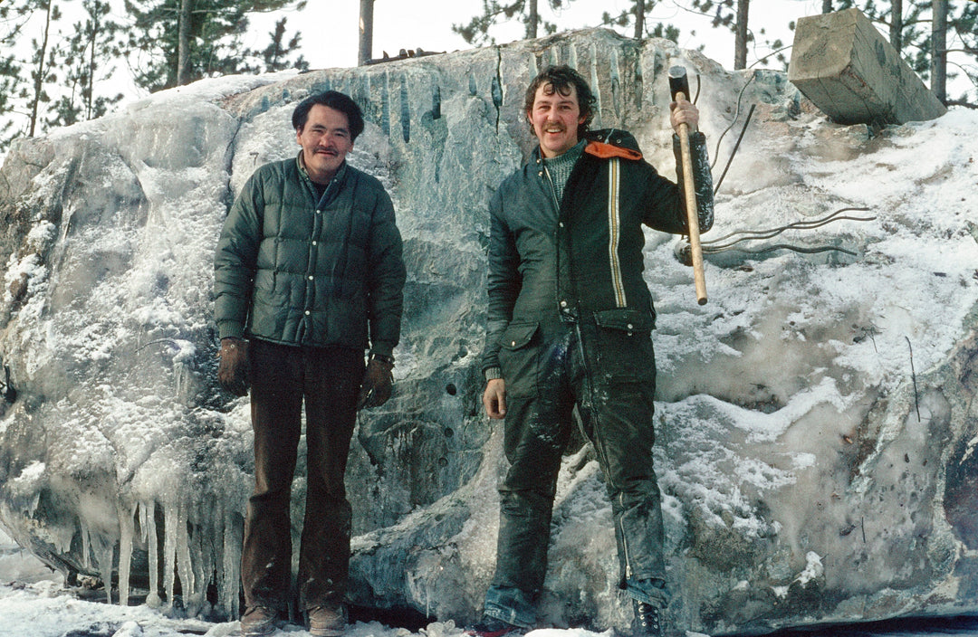 Two men in winter jackets pose beside a large icy rock at a jade mine, one holding a pickaxe.