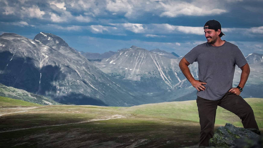 A man in a gray T shirt and cap stands on a grassy hill with towering mountains in the background, smiling.