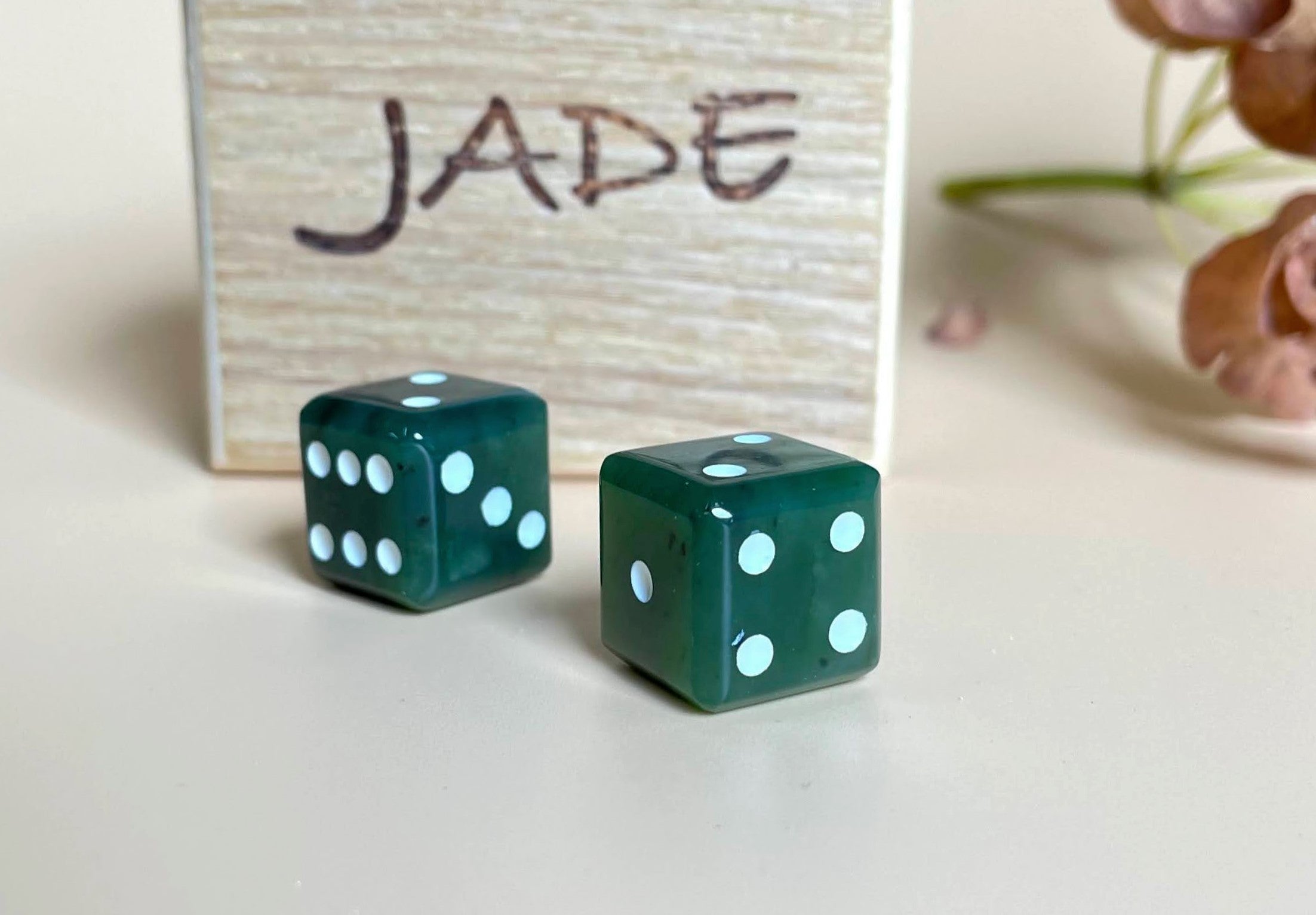 Two dark green jade dice with white pips on a white surface in front of a rustic wooden sign that reads Jade.