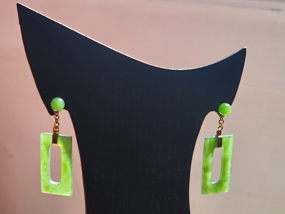 Two green jade rectangle drop earrings with gold connectors hang from a black display stand