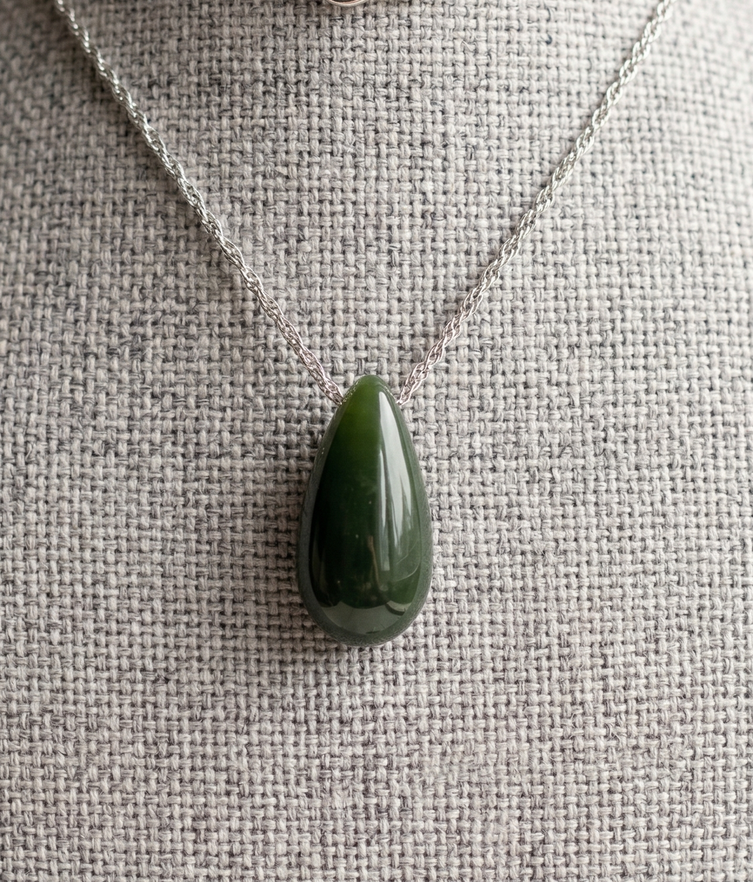 Dark green jade teardrop pendant on a silver chain displayed against a textured fabric background