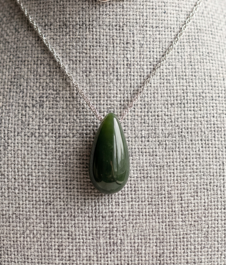 Dark green jade teardrop pendant on a silver chain displayed against a textured fabric background