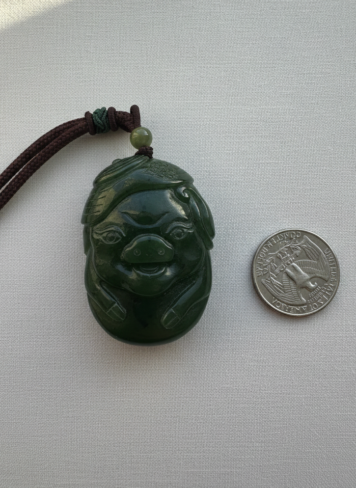 Hand carved green jade pig pendant on brown cord with a small bead, shown beside a coin for scale.