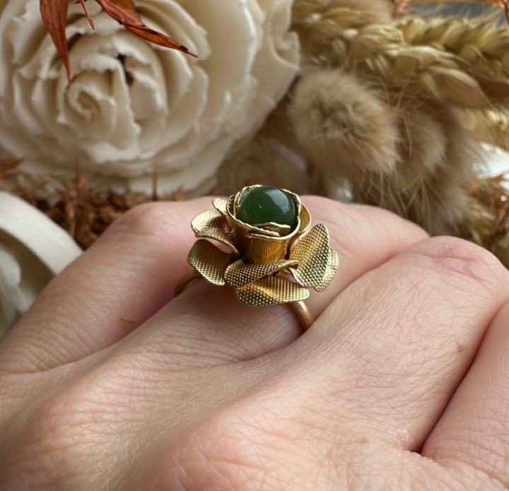 Gold ring with a green gemstone on a hand, surrounded by decorative flowers and leaves.