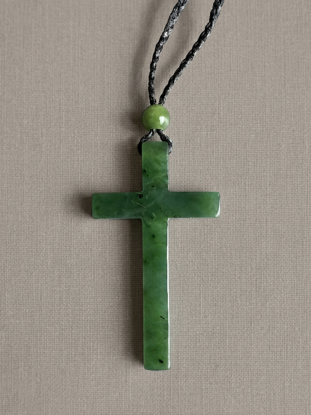 Green jade cross pendant on a braided black cord with a round green bead above it.