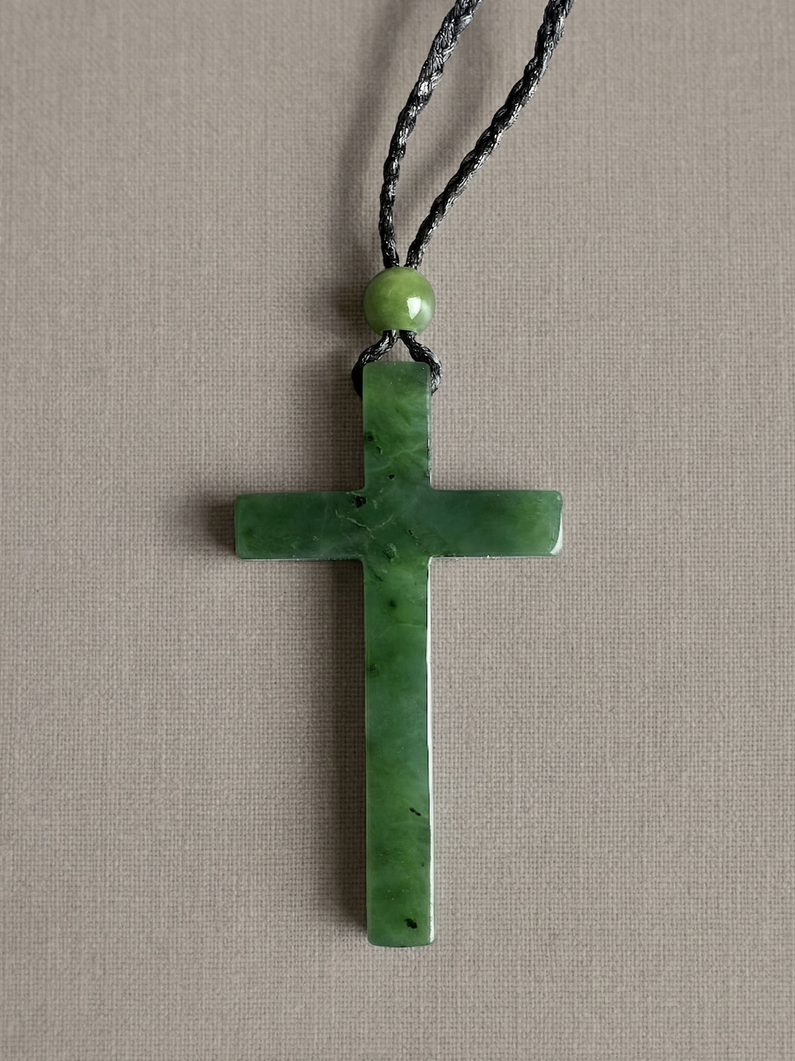 Green jade cross pendant on a braided black cord with a round green bead above it.