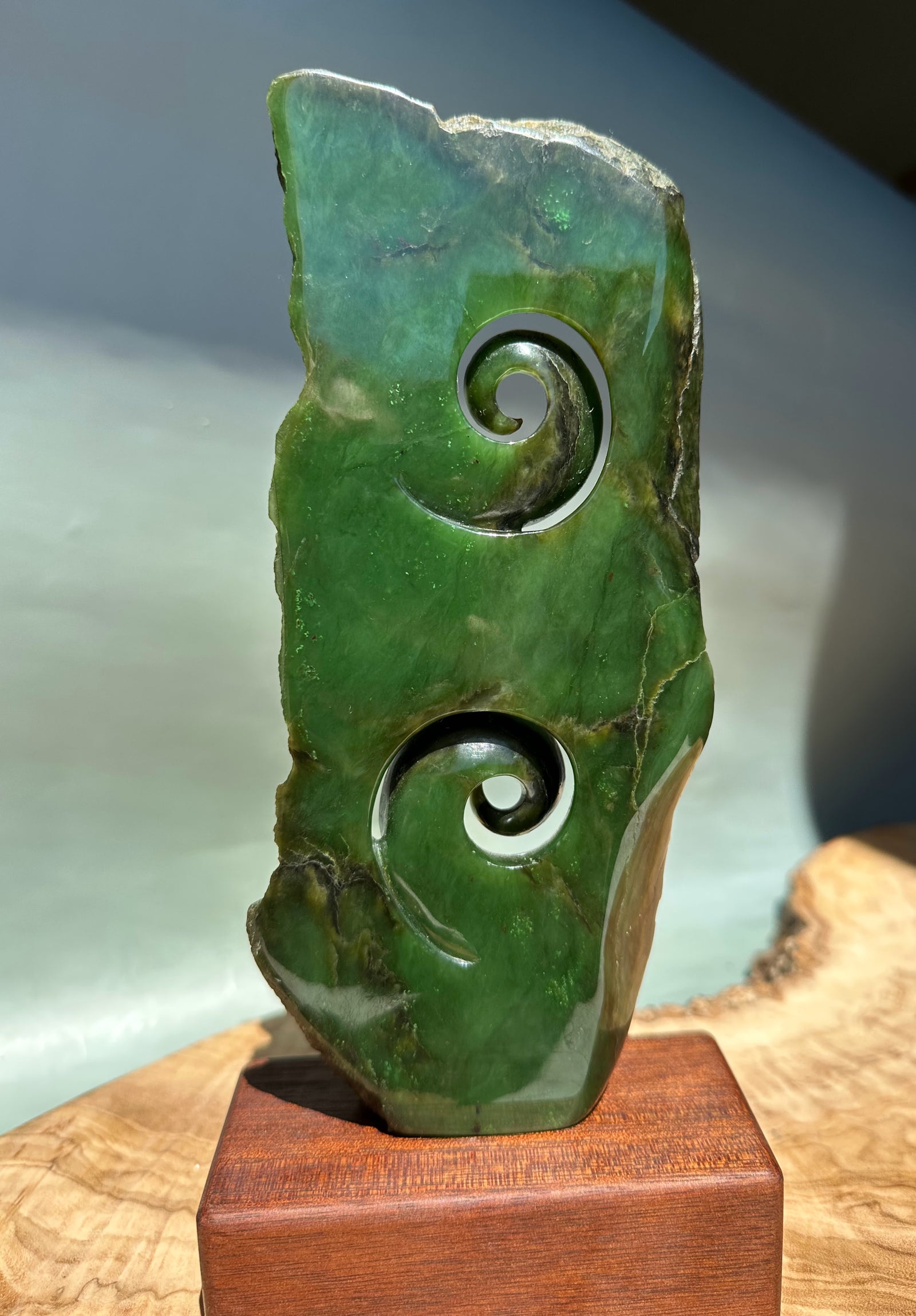 Green jade double koru sculpture carved in rock shown from the side with two curling spirals on a wooden pedestal