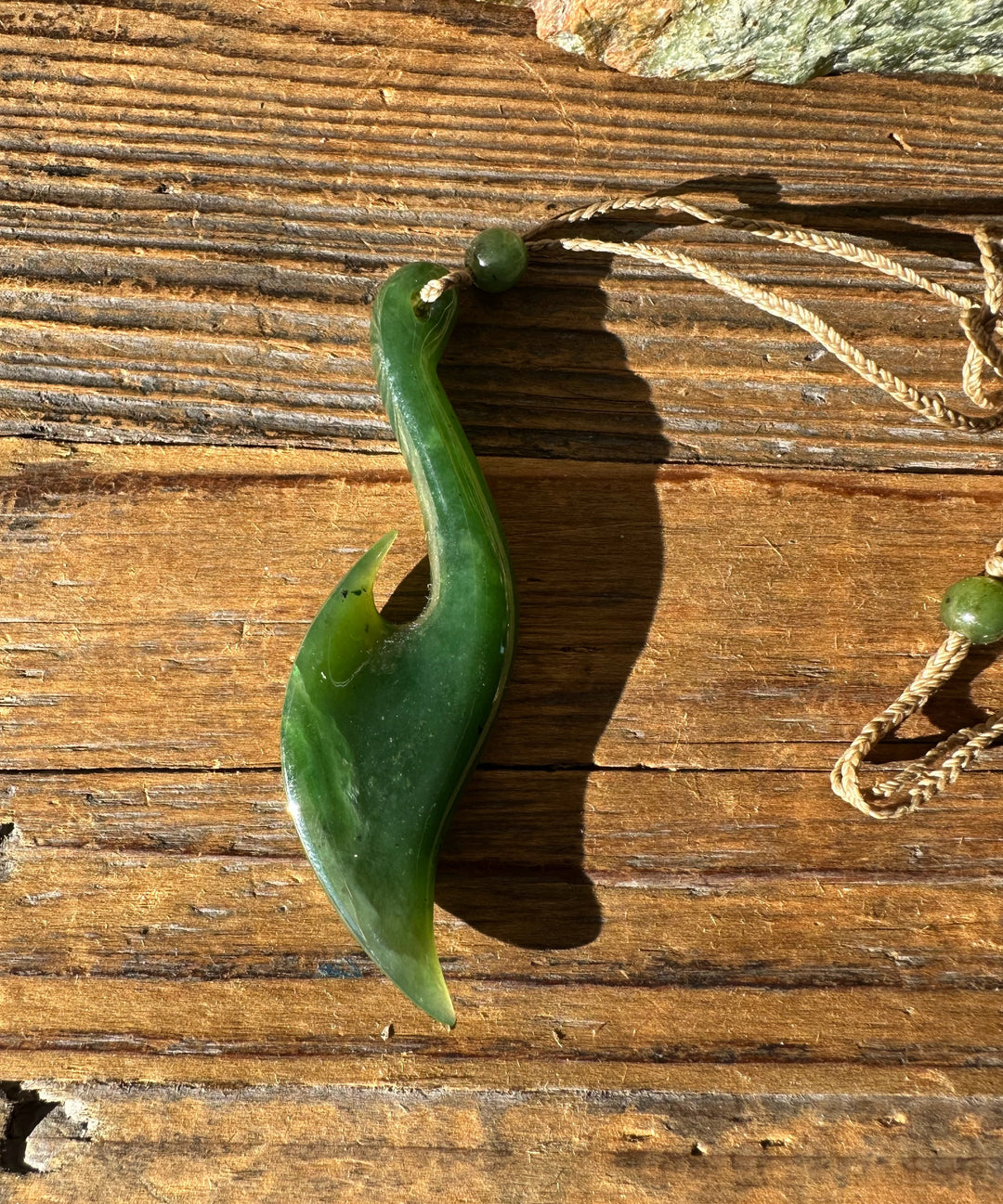 Jade fish hook pendant lying on a weathered wooden surface with a twine cord nearby