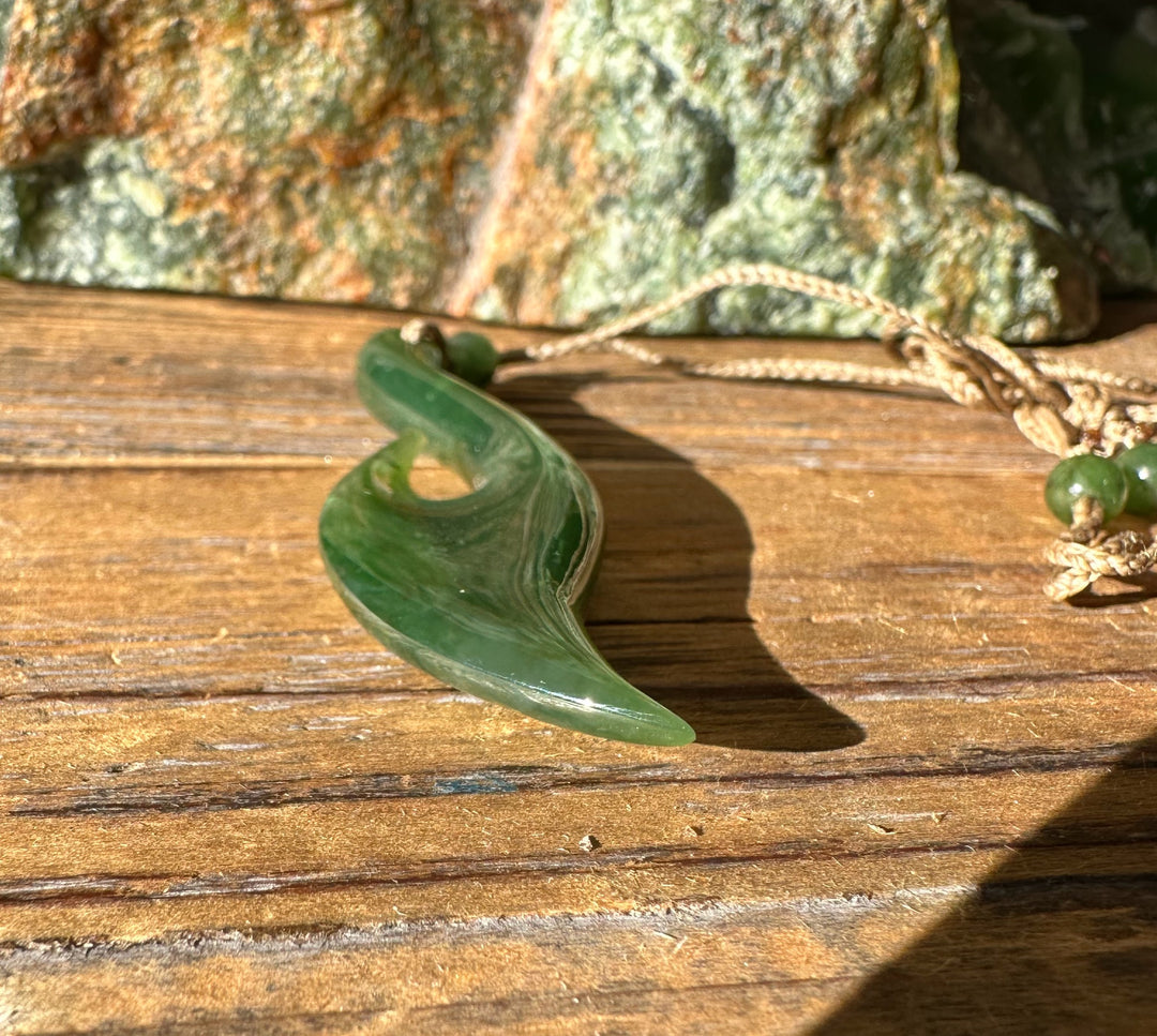 Jade fish hook pendant on rustic wood with knotted beige cord and green bead accents