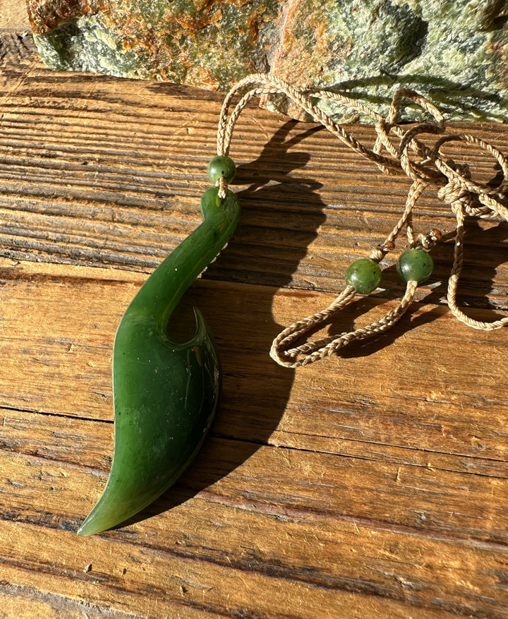 Close up of green jade fish hook pendant attached to braided cord with beads on a wooden surface