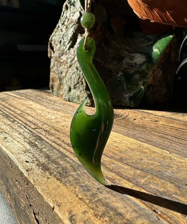 Jade fish hook pendant on a twine cord with a small green bead, set against a rock backdrop