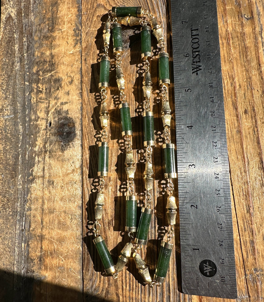Vintage jade necklace with long green cylindrical beads and gold caps laid on a wooden surface beside a ruler for scale