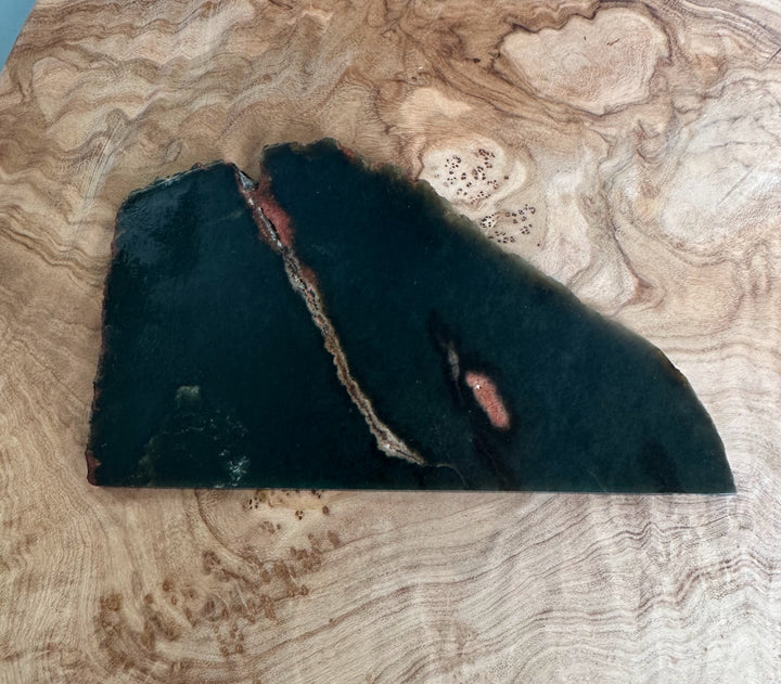 Wyoming Jade Slab 105g dark green with a diagonal rust vein and reddish patches on a wood surface.