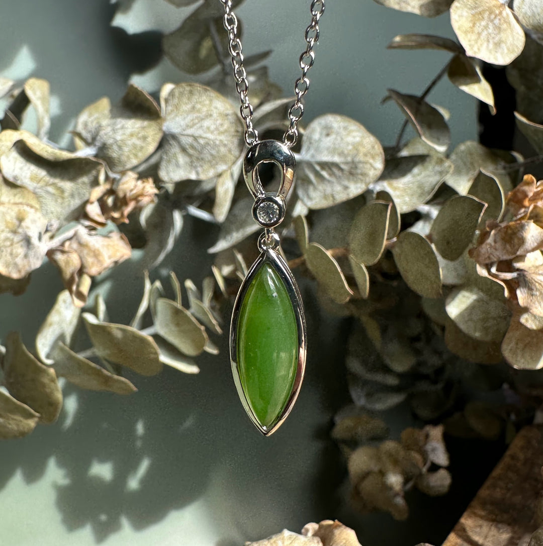 Close-up of a green jade marquise pendant in a silver bezel on a chain with a botanical backdrop