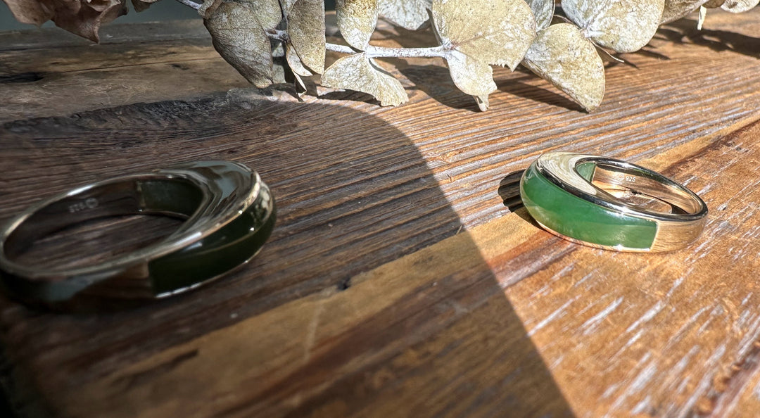Two Canadian jade rings with green inlays and silver bands rest on rustic wood beside dried leaves