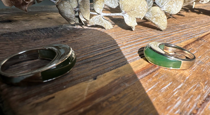 Two Canadian jade rings with green inlays and silver bands rest on rustic wood beside dried leaves