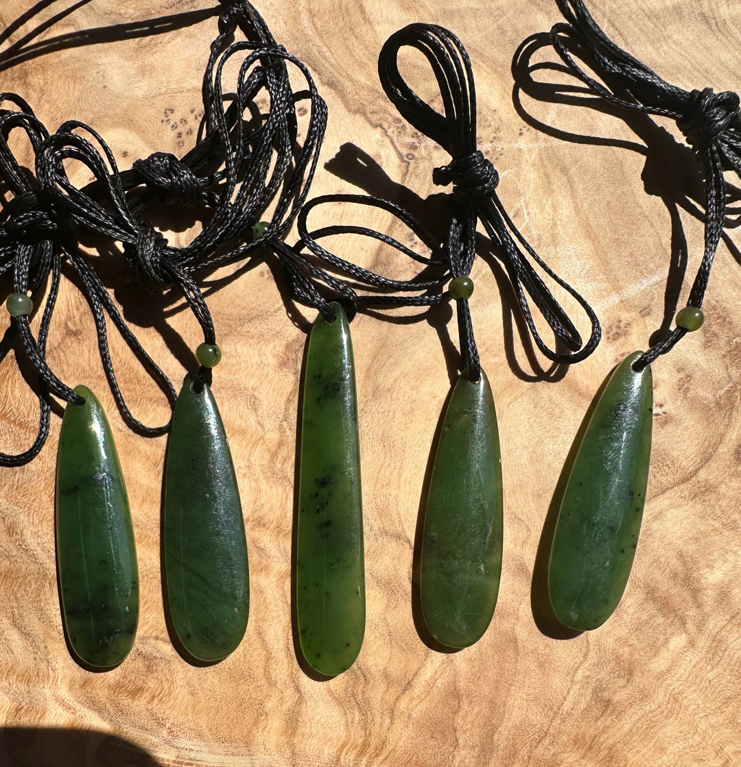 Five green jade drop pendants with black cords laid on a wooden surface