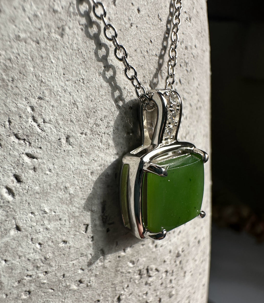 Side-angle view of the green jade pendant with silver frame and CZ bail resting on a textured stone surface