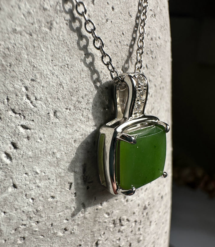 Side-angle view of the green jade pendant with silver frame and CZ bail resting on a textured stone surface