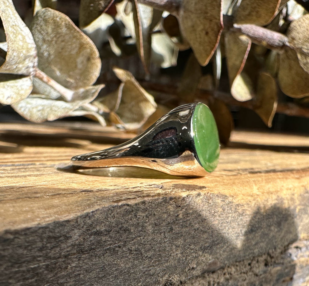 Angled view of a silver ring with a green jade oval on a wooden surface with dried leaves in the background.