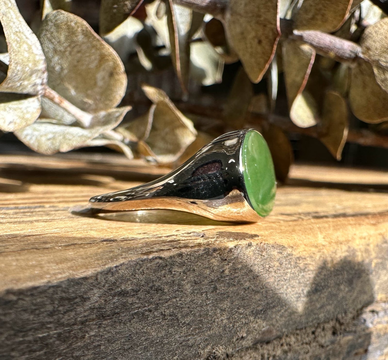Angled view of a silver ring with a green jade oval on a wooden surface with dried leaves in the background.