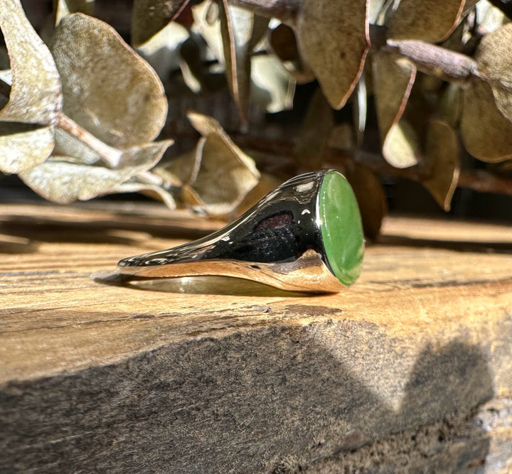 Angled view of a silver ring with a green jade oval on a wooden surface with dried leaves in the background.