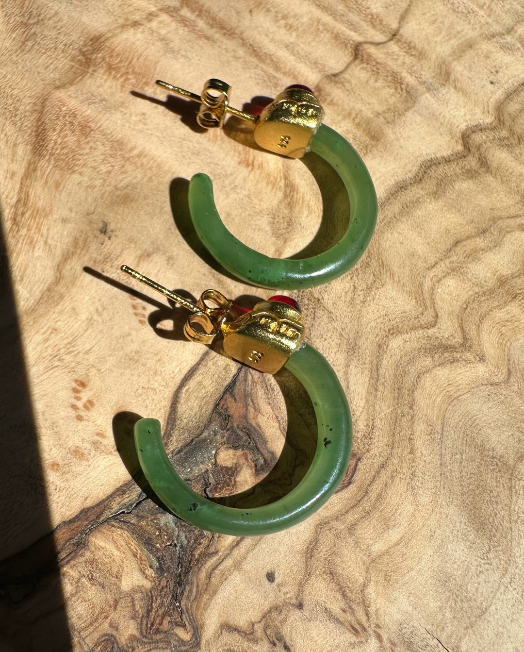 Pair of green jade hoop earrings with gold caps and red cabochons on a tan textured fabric surface.