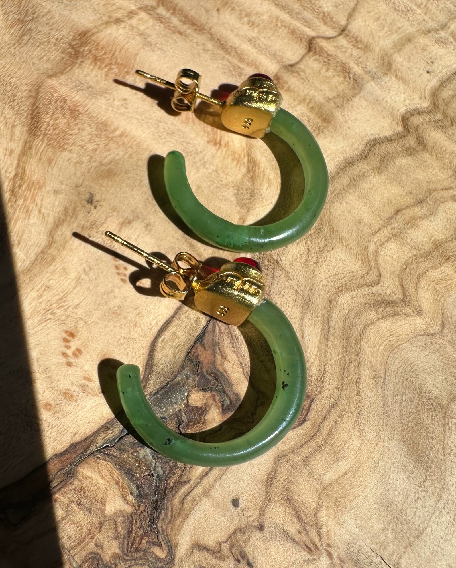 Pair of green jade hoop earrings with gold caps and red cabochons on a tan textured fabric surface.