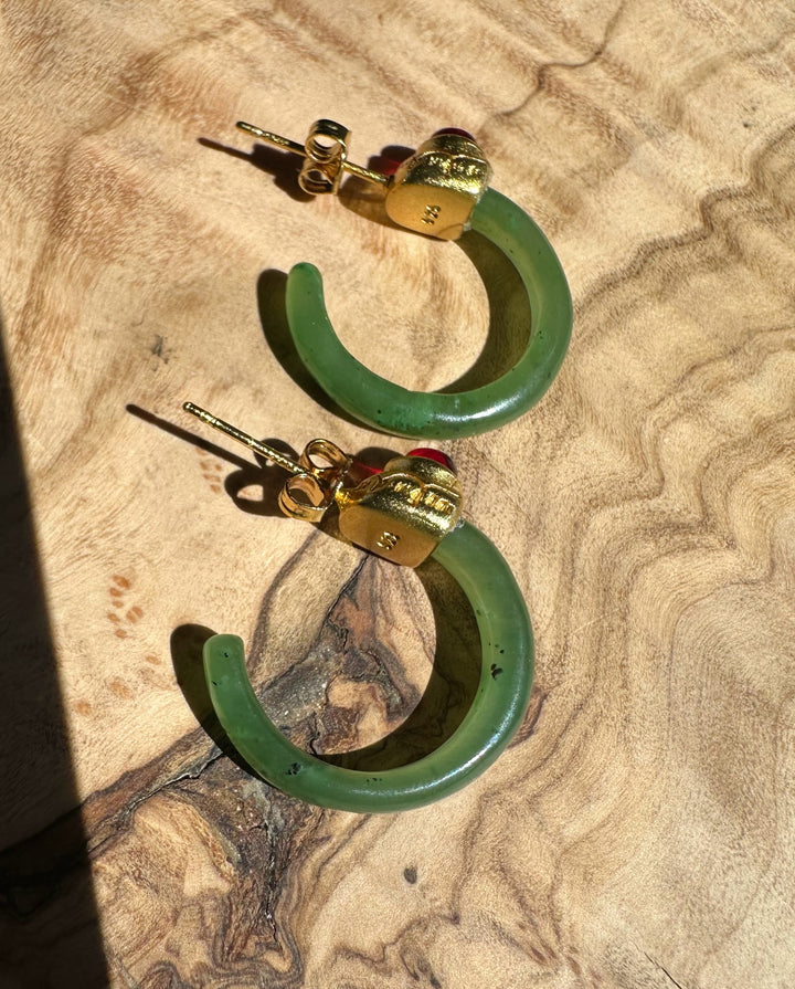 Pair of green jade hoop earrings with gold caps and red cabochons on a tan textured fabric surface.