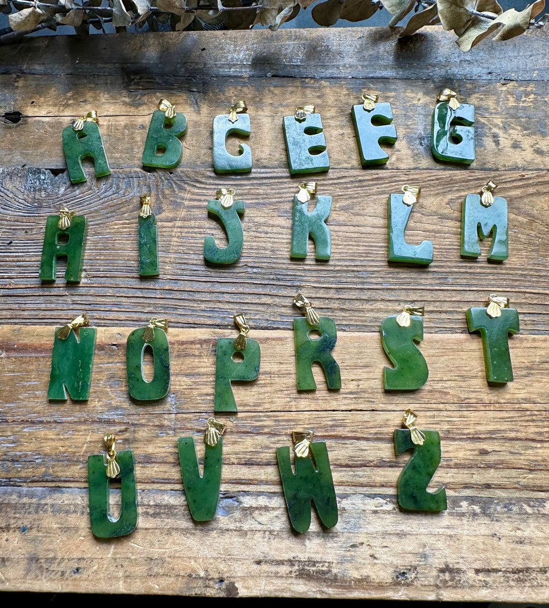 Vintage green jade letter pendants with gold bails arranged on a rustic wooden surface