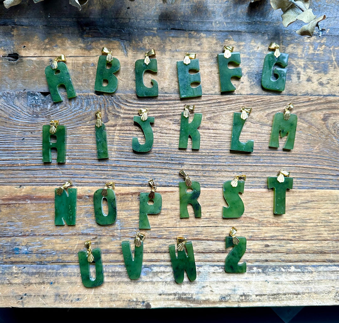 Green jade alphabet pendants with gold loops laid out on weathered wood