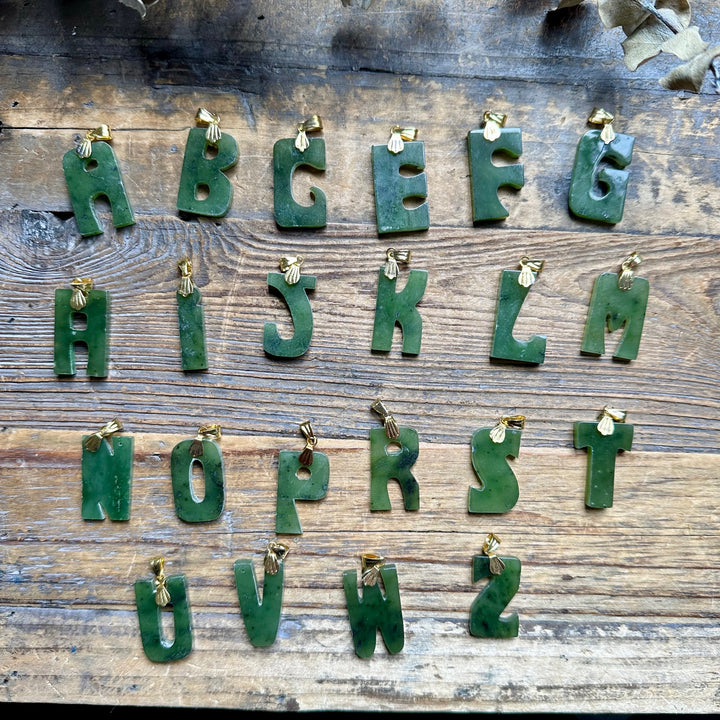 Vintage jade letter pendants with gold bails laid out on a wooden background