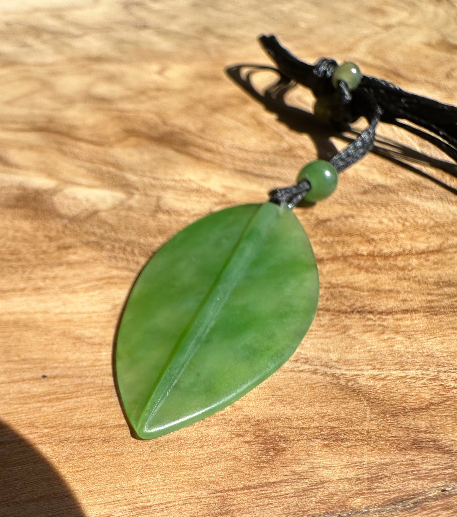 Siberian Jade Leaf Pendant on black cord with a green bead resting on a wooden surface