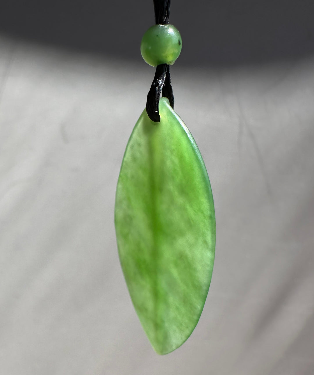 Siberian Jade Leaf Pendant close up with a green bead on braided black cord against a blurred background