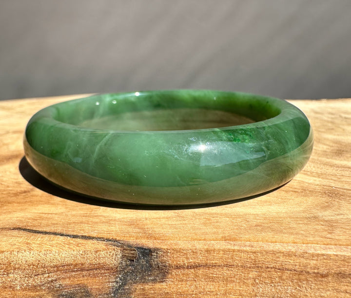 Green jade bangle with darker natural inclusions on a wooden table, captured in a close up view