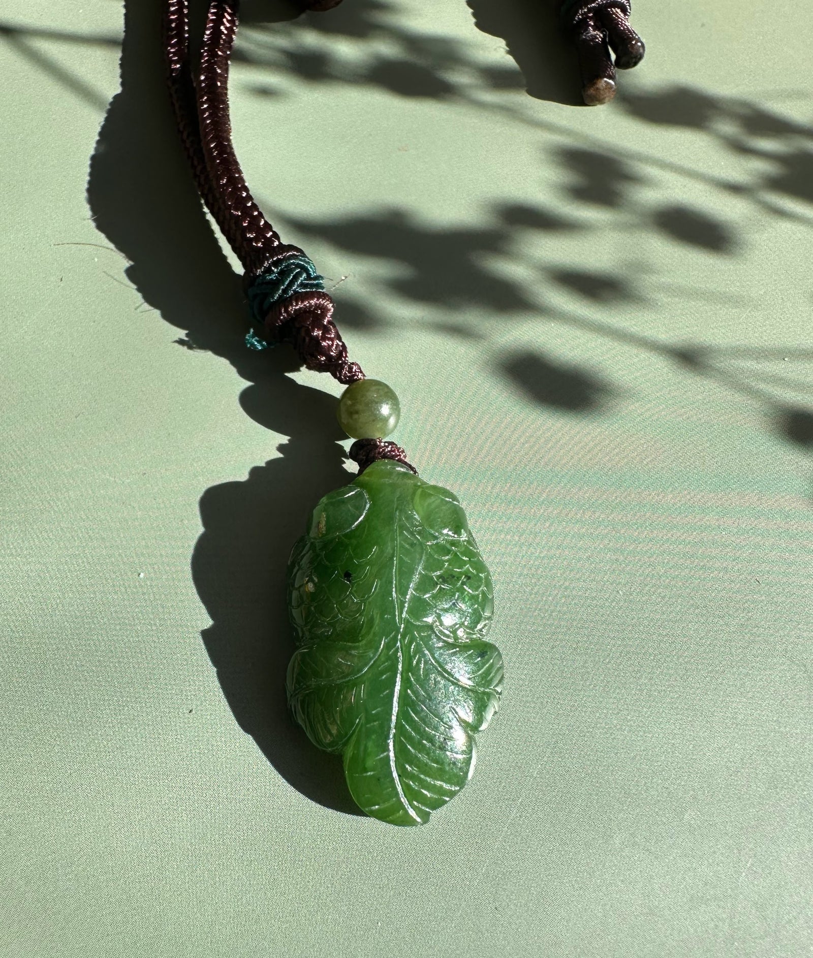 Green jade goldfish pendant carved with scales, attached to a brown cord with a pale bead.