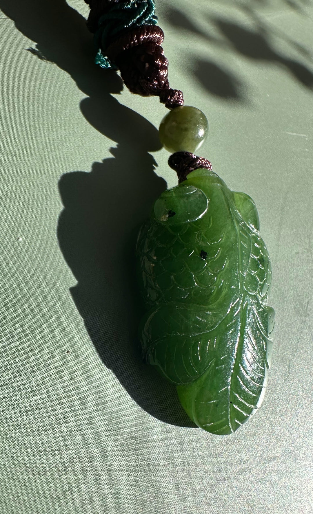Close-up of green jade leaf pendant on braided cord with bead against a pale background