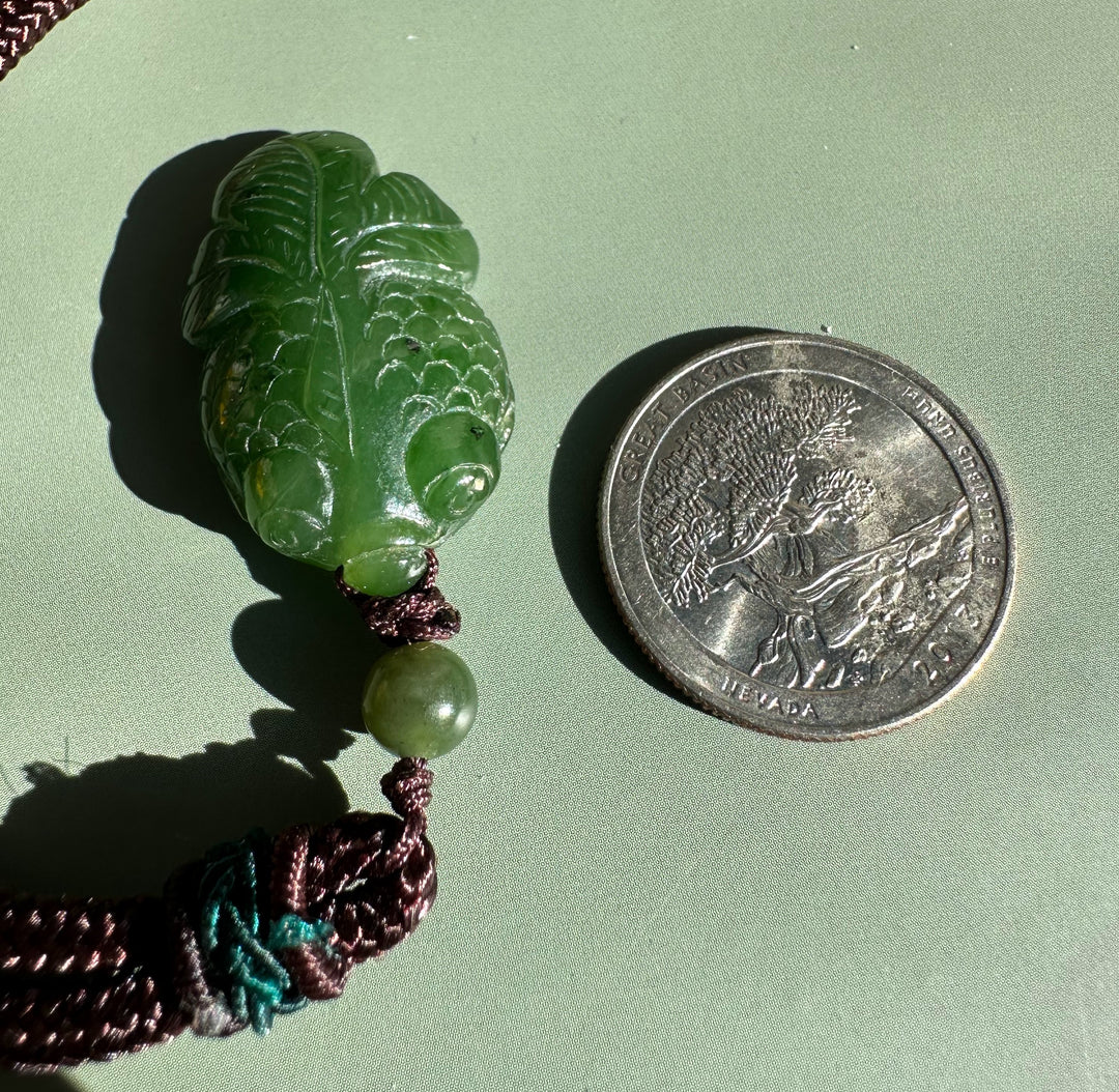 Leaf shaped green jade pendant with prominent veins on a brown cord beside a coin for scale.