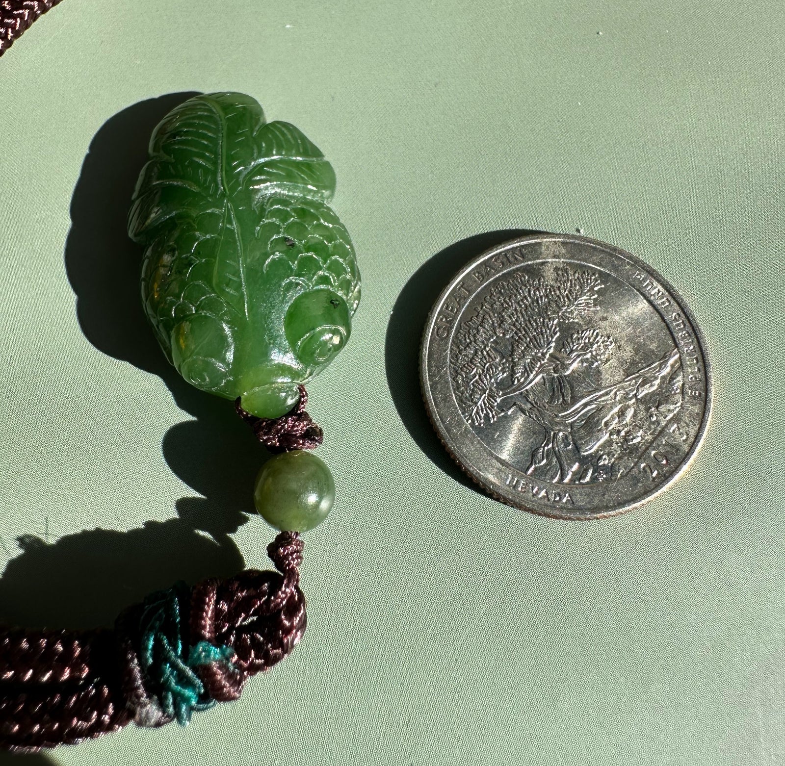 Leaf shaped green jade pendant with prominent veins on a brown cord beside a coin for scale.
