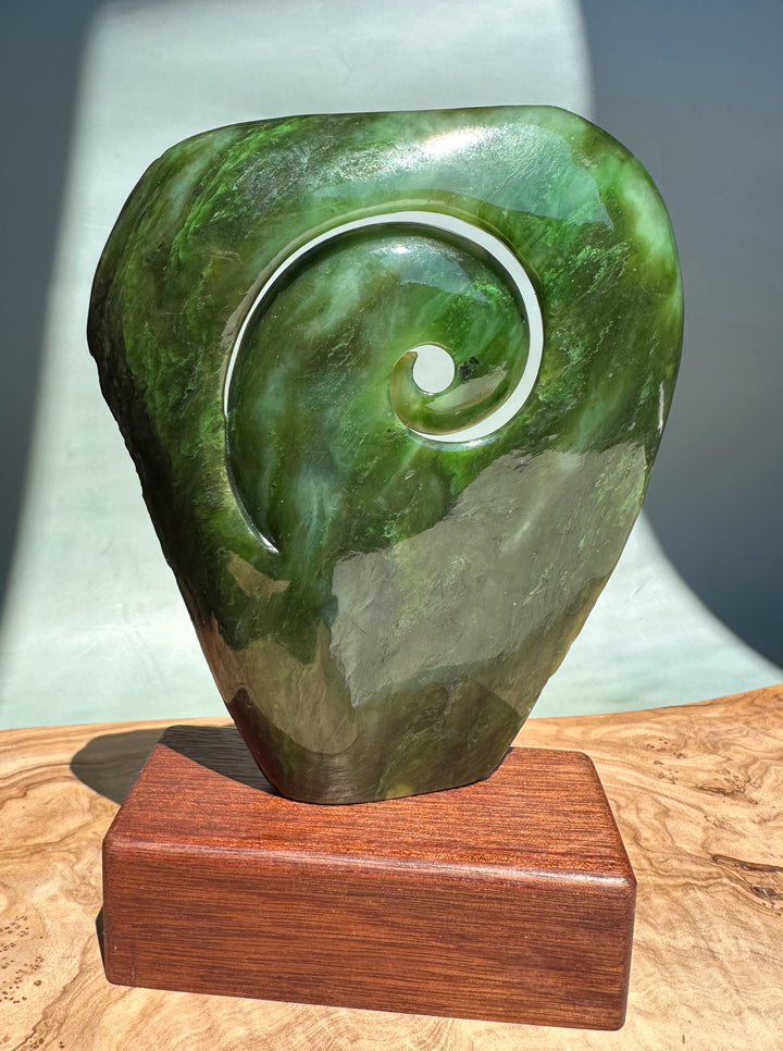 Green polar jade koru sculpture with a white spiral accent carved into the stone, mounted on a wooden base.