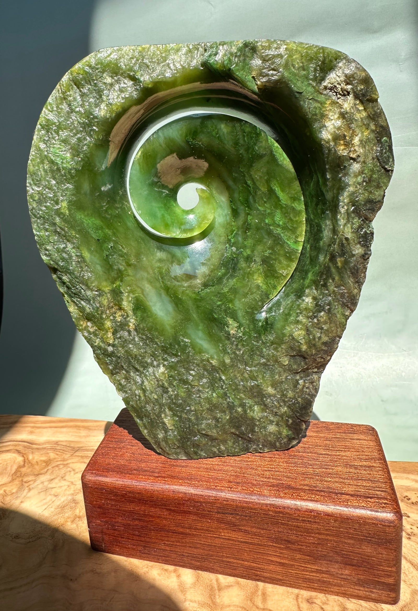 Polar Jade Koru Sculpture set in rough green stone with a carved spiral center on a wooden pedestal
