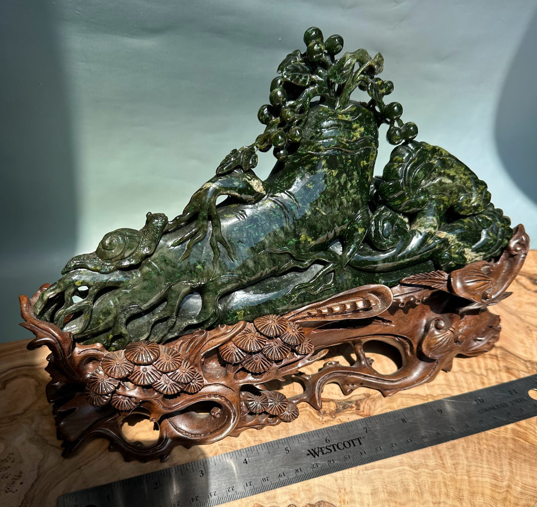 Green jade Cassiar sculpture of a reclining mythical beast with a vine tree on its back, set on a carved wooden stand.