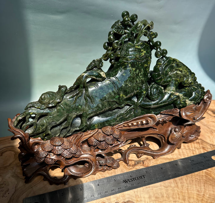 Green jade Cassiar sculpture of a reclining mythical beast with a vine tree on its back, set on a carved wooden stand.