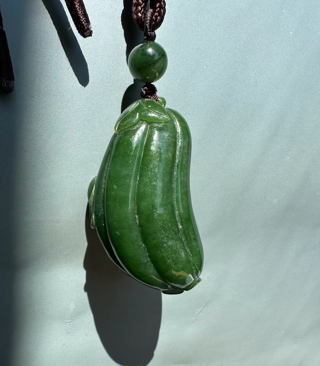 Side view of green jade gourd pendant with leaf cap and round bead on a dark braided cord
