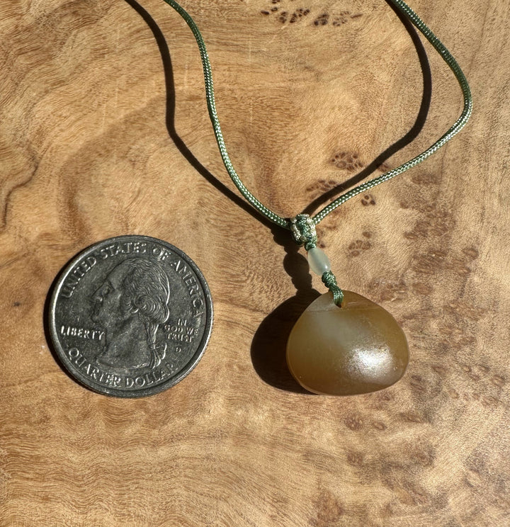 Dainty honey nephrite jade pendant on a green braided cord with a pale jade bead, placed on wood beside coins.