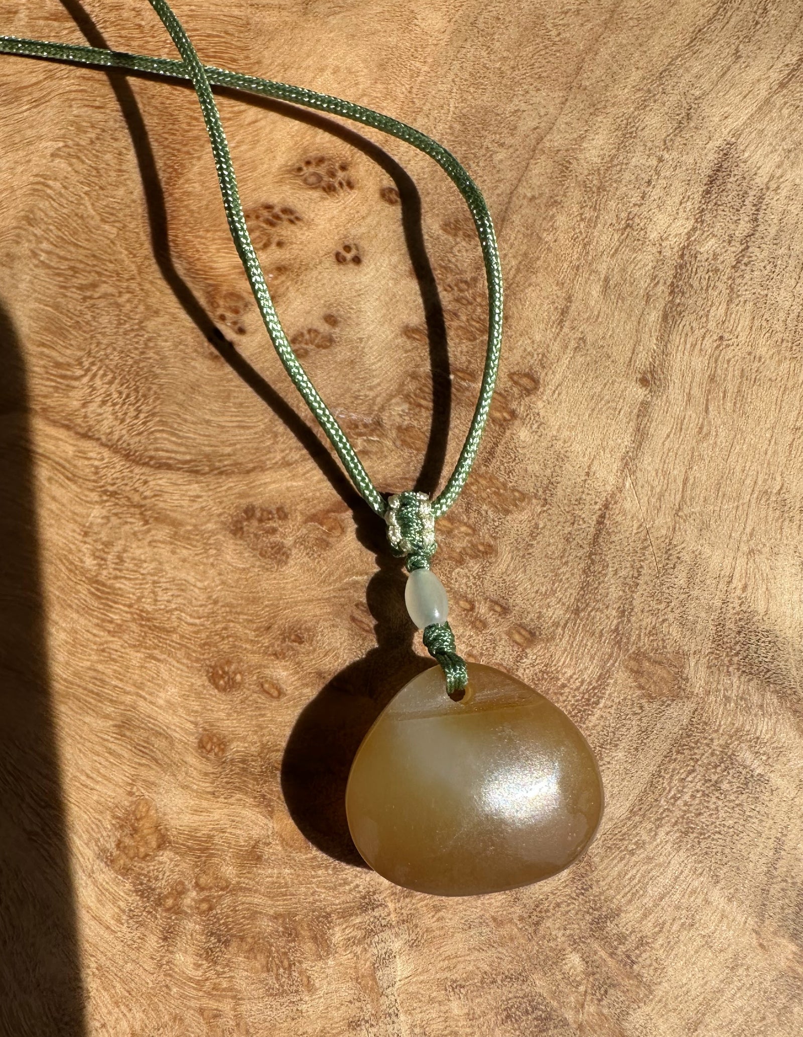 Dainty honey nephrite jade circular pendant on a green braided cord with knot on a wood surface