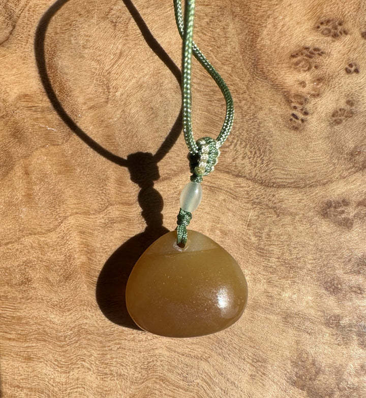 Jade pendant on a green braided cord with a pale oval bead above a brown stone pendant against a wooden surface.
