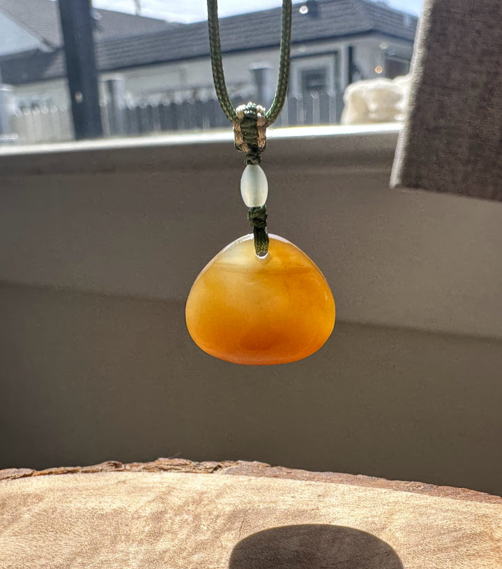 Pale jade bead and orange teardrop pendant on a green braided cord with knot against a neutral backdrop