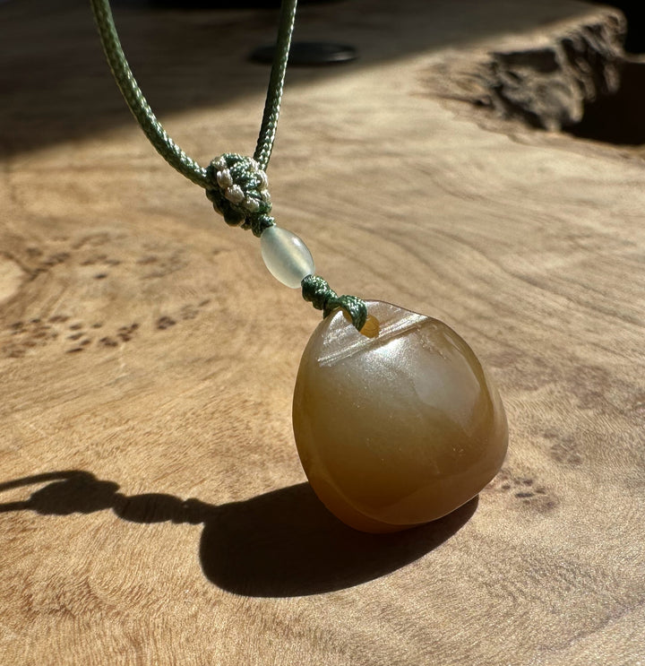 Close-up of a knotted green cord holding a pale nephrite jade pendant on a textured wooden surface.