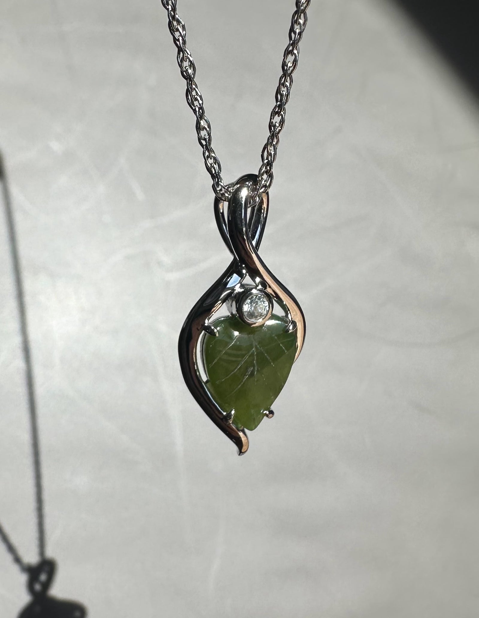 Close up of the pendant on a chain showing the green jade leaf and silver infinity curves against a pale background